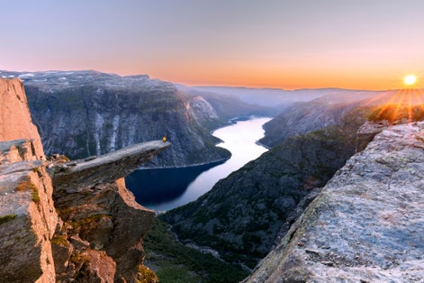 A man sits while throwing his arms in the air on the mountain's cliff edge of Trolltunga throning over Ringedalsvatnet watching the sunset in the snowy Norwegian mountains near Odda, Rogaland, Norway