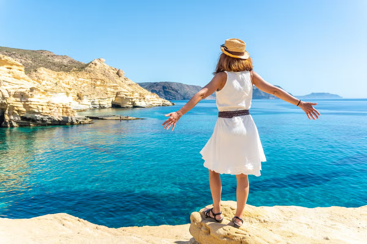 A woman in a white dress and straw hat stands on a rocky cliff in Rodalquilar, Cabo de Gata, overlooking the turquoise sea..png