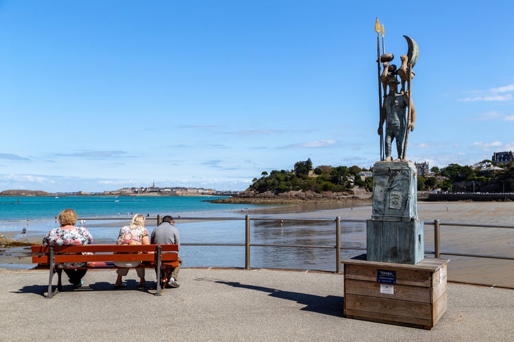 People rest on the beach promenade at the Man with Birds monument (by Tomek) at low tide.