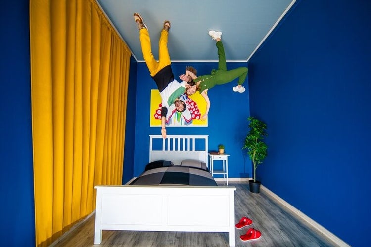 An optical illusion room with two people appearing to hang upside-down, featuring blue walls, yellow curtains, and colorful decor..jpg