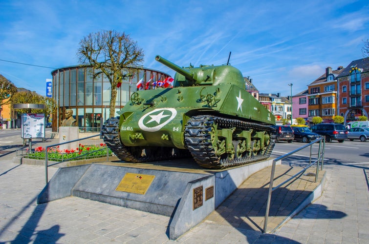 Photo of Sherman tank in original livery freshly painted on stone plinth. Information plaque to front. Alongside statue of American General McAuliffe in flowered enclosure, Bastogne Belgium.