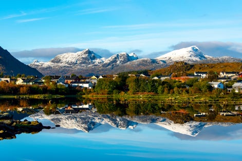 Snow-covered mountains reflect in the still fjord waters near Leknes, Norway, with a small village of colorful houses nestled at the base.