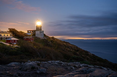 photo of Kullen lighthouse shining its light after sunset. Denmark and Sweden in the background.