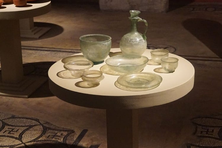 A round display table showcases ancient glass bowls, plates, and a jug in a dimly lit museum setting..jpg