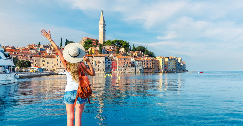 Photo of young tourist woman on harbour holding her hand up wave to beautiful Rovinj city, Croatia.