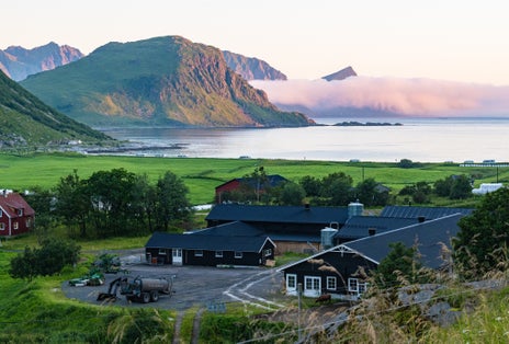 A rural farmstead near Leknes, Norway, surrounded by green fields and steep coastal hills, with soft evening light and fog rolling over the sea.