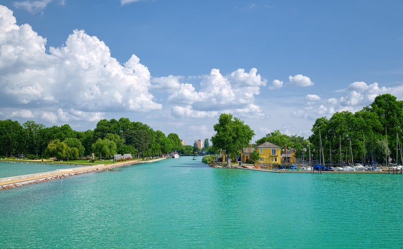 photo of view of View to entrance to Siofok harbor at Balaton lake, Hungary, with crystal clear water of emerald color, green coastline, puffy clouds on sky.