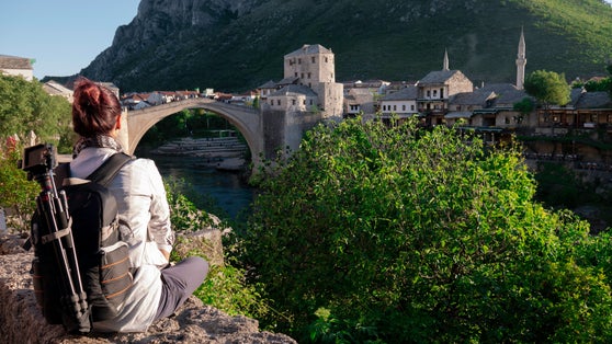 Photo of tourist  woman sitting on a stone wall and looking at the Old bridge (Stari most) in Mostar, Bosnia and Herzegovina.