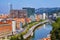 Photo of aerial view of Bilbao, Spain city downtown with a Nevion River, Zubizuri Bridge and promenade. Mountain at the background, with clear blue sky.