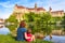  PHOTO OF VIEW OFFamily sit near medieval Sigmaringen Castle, Germany. Young woman and her baby are by river in summer. Scenic view of old famous castle on cliff on sunny day. Travel and vacation in Baden-