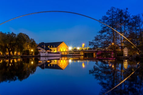 photo of Stangan or Kinda kanal at twilight time. Yellow lights shine on the Scandinavia building contrast with the blue sky. The metal curve structure reflects on the still water surface. Linköping, Sweden.