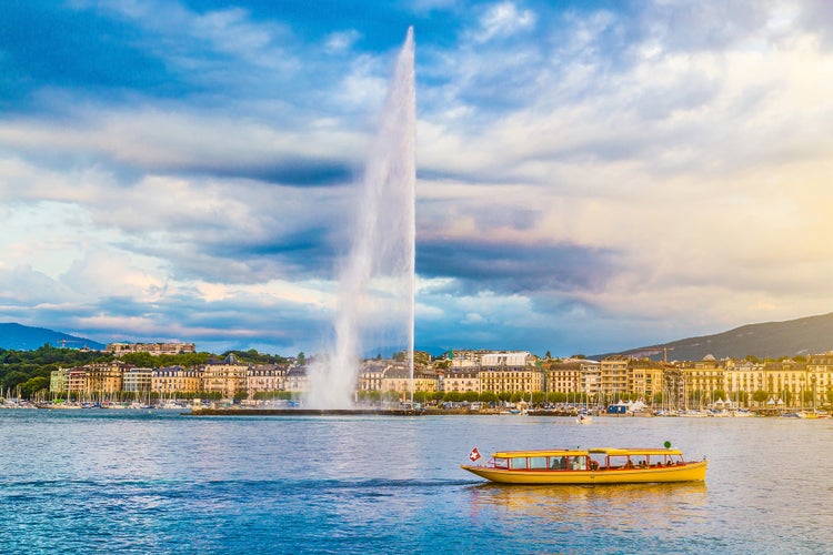 Photo of panoramic view of Geneva skyline with famous Jet d'Eau fountain and traditional boat at harbor district in beautiful evening light at sunset, Canton of Geneva, Switzerland.