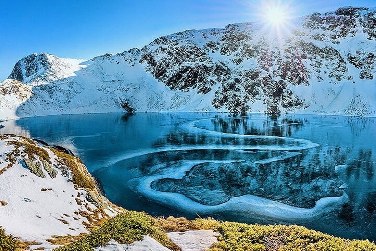 A frozen alpine lake surrounded by snow-covered mountains under a bright blue sky, with the sun shining brightly, creating an icy pattern..jpg