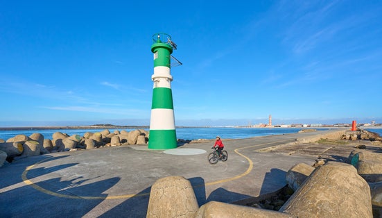 happy senior woman cycling below the navigation sign and ligthouse of Barra at the harbor entrance of Aveiro, Portugal