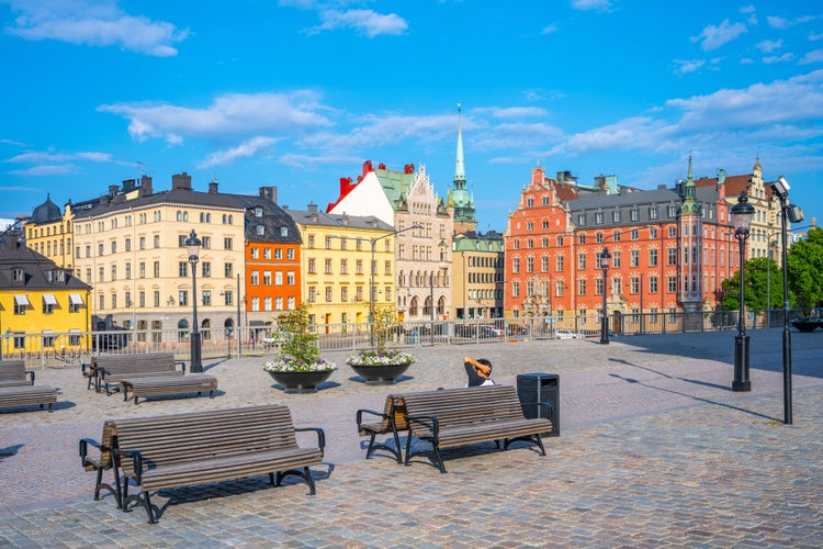 Benches line a cobblestone square in Stockholm bathed in sunlight, surrounded by historic colorful buildings under a clear blue sky. Sweden