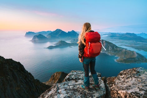 A female hiker with a red backpack stands on a rocky summit in Ryten, overlooking dramatic sea cliffs and islands in Norway’s Lofoten archipelago at sunset.