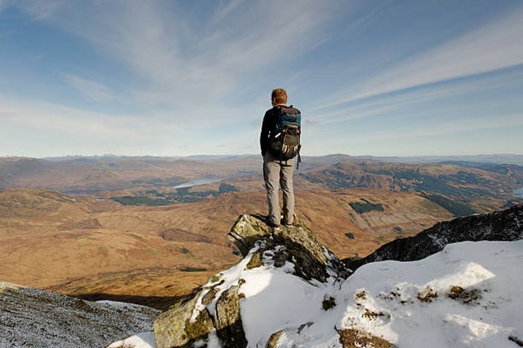 A man standing at the summit of Ben Lomond, admiring the expansive view of Loch Lomond and the surrounding Scottish Highlands.jpg