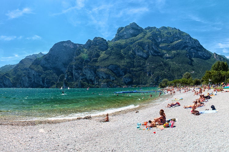 Beach with sunbathing people at lake in mountains, cliffs and blue sky in background, Lago di Garda, Riva del Garda, Italy