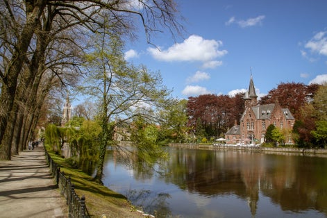 Photo of tourists going for a walk at the Minnewater lake in the Minnewaterpark. On the shores of the lake is the Greenhouse and the tower of the Church of Our Lady in the background.