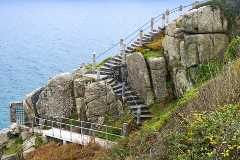 Photo of Steep steps descending between rocks and cliff with handrails beside the beautiful blue ocean at Minack Theatre near Porthcurno Beach, Cornwall, England.