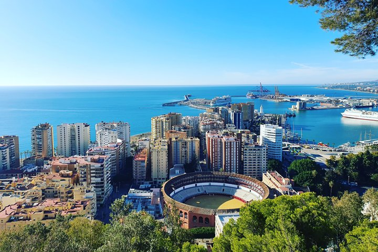 A panoramic view of a city featuring a stadium, a large body of water, and the historic Gibralfaro Castle in the background..png