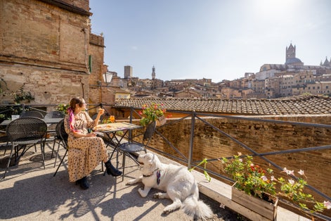 photo of view ofYoung woman having lunch with pizza and wine, sitting with dog at outdoor restaurant with beautiful view on the old town of Siena. Concept of italian cuisine and travel