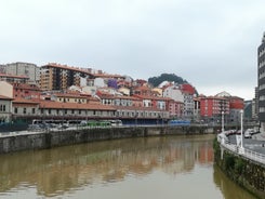 Photo of aerial view of Bilbao, Spain city downtown with a Nevion River, Zubizuri Bridge and promenade. Mountain at the background, with clear blue sky.