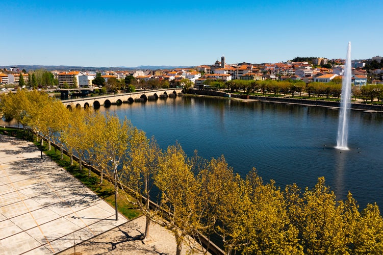 Photo of scenic view of Mirandela cityscape on banks of Tua River with landscaped embankment, modern water fountain and ancient stone arched bridge across river on sunny spring day, Portugal.