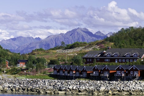 photo of view of Homes along shore protected by stone barrier, with mountains behind in Stokmarknes, Norway