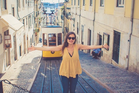 Photo of Happy woman with open arms and sunglasses in Lisbon, Portugal, Europe enjoying holidays in front of Bica Funicular. Elevador or Ascensor da Bica is a popular tourist attraction. Yellow vintage tram.