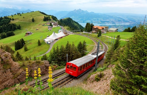 photo of a sightseeing train traveling on the cogwheel railway through green grassy meadows on Mt. Rigi, with rugged Pilatus peaks among majestic mountains in background on a cloudy summer day in Switzerland.