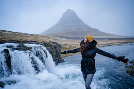 photo of Happy free beautiful girl dancing with open arms of happiness in the wind Iceland nature. In background on famous Kirkjufell Mountain with colorful, dramatic sky.Europe travel tourist enjoying wint