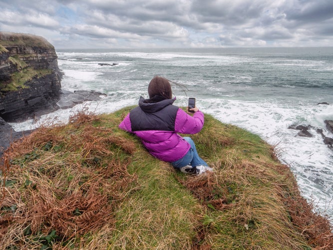 photo of view of Teenager tourist girl laying on edge of a cliff and looking at stunning nature scene with rough stone coastline with cliff, ocean waves and dramatic sky. Mullaghmore, county Sligo, Ireland.