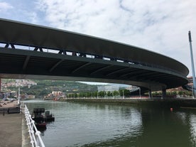 Photo of aerial view of Bilbao, Spain city downtown with a Nevion River, Zubizuri Bridge and promenade. Mountain at the background, with clear blue sky.