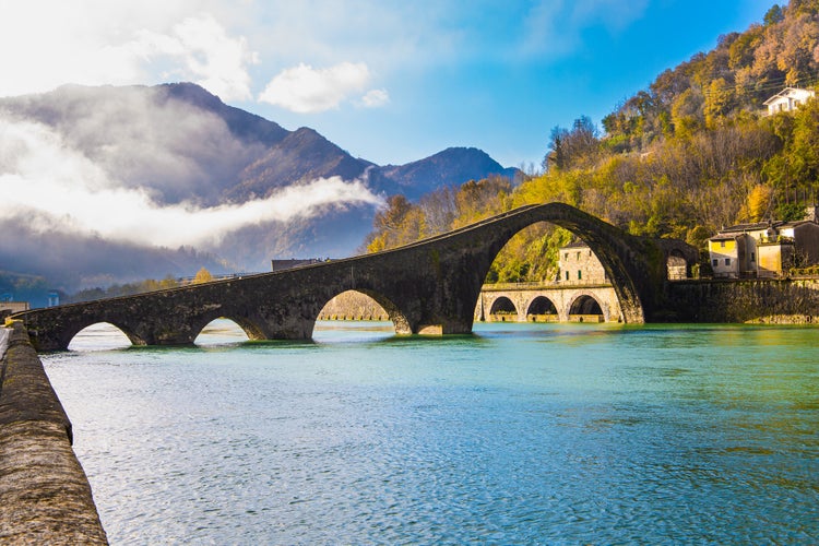 photo of view of Italy, province of Lucca. Fancy Medieval Bridge - Devil's Bridge crosses the Serchio River. The green emerald cold water of the river reflects the ancient asymmetrical arches of the bridge