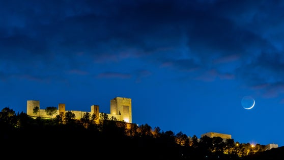 photo of Castillo de Santa Catalina in Jaén, Spain. illuminated at night, with a cloudy blue sky, and the waxing crescent moon on the right side of the image.