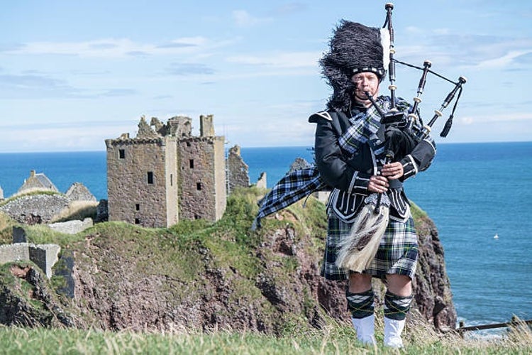 A bagpiper dressed in full traditional Scottish attire with Dunnottar Castle in the background.jpg