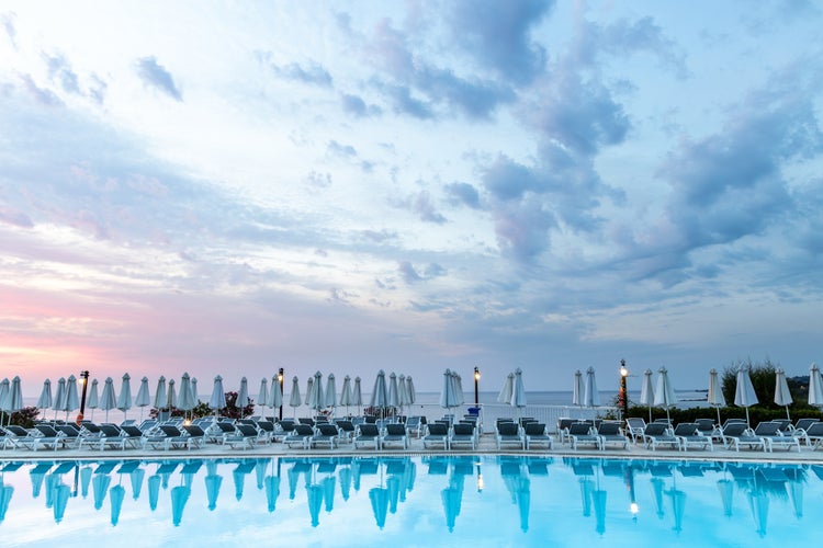 Photo of Aegean sea sunrise behind pool, umbrellas and sun deck chairs at Kallithea, Greece.