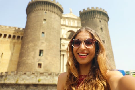 photo of Smiling hipster woman with sunglasses taking selfie photo in Naples with Castel Nuovo castle on the background, Naples, Italy.