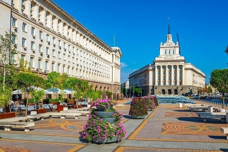 A city square with flower planters, benches, and trees. Historic buildings with ornate facades surround the area under a clear blue sky..jpg