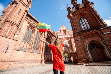 photo of young female tourist with photo camera and lithuanian flag standing back near the famous gothic church in the old town of Vilnius. Woman having great vacations in Lithuania.